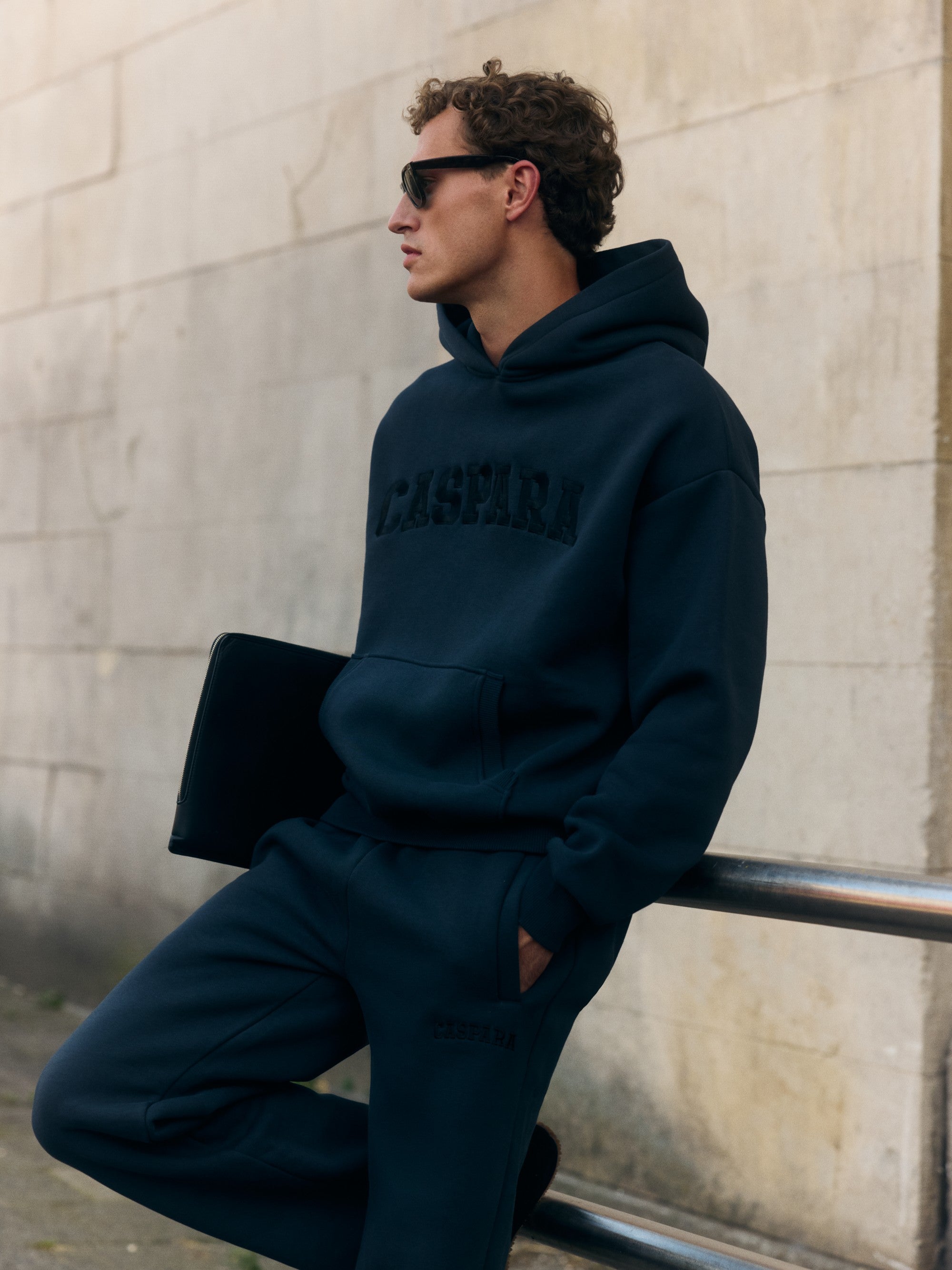 Man wearing a black CASPARA logo hoodie and sunglasses outdoors, leaning against a railing.